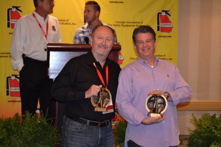 Two men holding trophies in front of a yellow wall with fire extinguishers on it