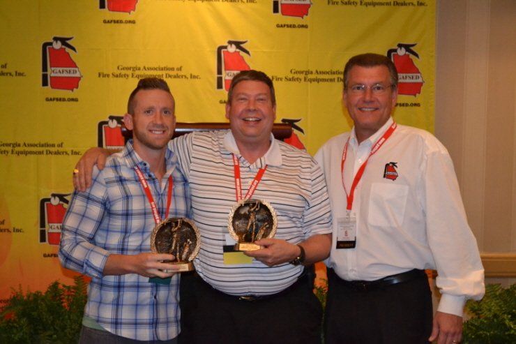 Three men holding trophies in front of a fire extinguisher wall