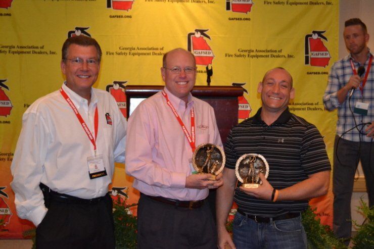 Three men holding trophies in front of a fire extinguisher wall