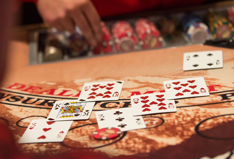 Blackjack table: Dealer's hand reaching for cards; player cards visible: Queen, 5, 10, 7, 7, 6; chips present.