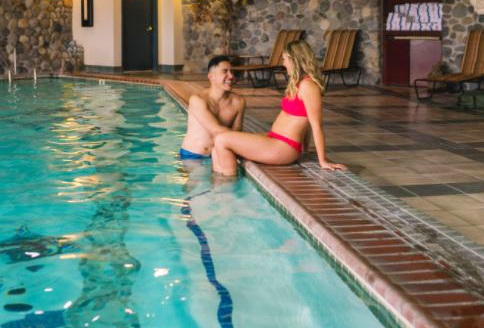 Couple in a pool; woman sits on edge in red bikini, man in water, smiling. Indoor pool, stone walls.