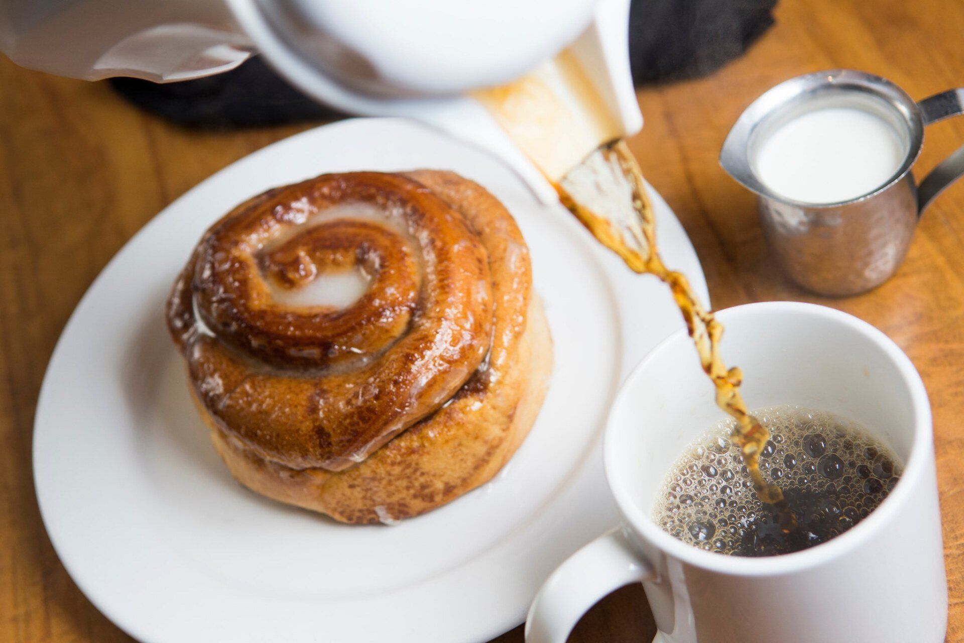 Coffee being poured into a white mug next to a cinnamon roll on a plate and a small creamer pitcher.