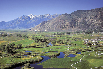 Green valley with golf course and ponds, mountains in the background, blue sky.