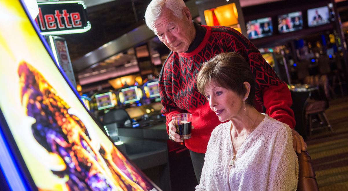 An older man and woman at a slot machine in a casino. The man is looking over the woman's shoulder.