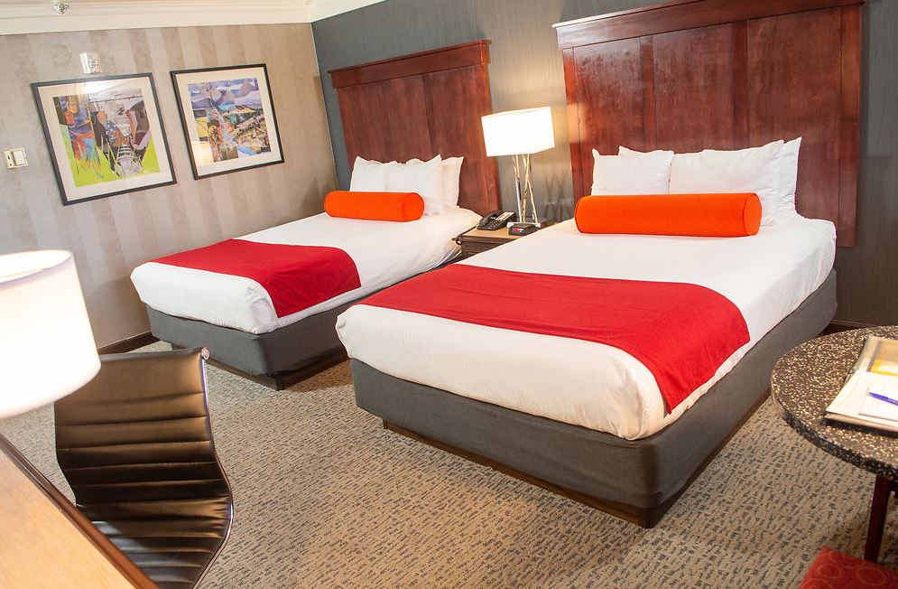 Two beds in a hotel room with red accents, lamps, artwork, and a small table.