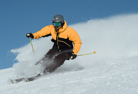 Skier carves down a snowy slope, kicking up a white spray. He wears a yellow jacket, black pants, and a helmet.