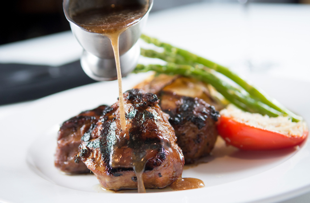 Grilled meat with gravy being poured on it, asparagus, and a tomato half on a white plate.