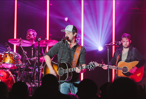 Band on stage performing under pink lights; guitarist sings and plays, drummer in back.