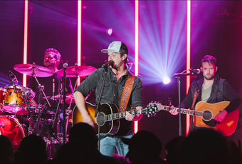 Band on stage performing under pink lights; guitarist sings and plays, drummer in back.