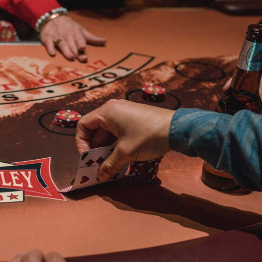 Person holding cards at a blackjack table; beer bottle nearby, red and brown tones.
