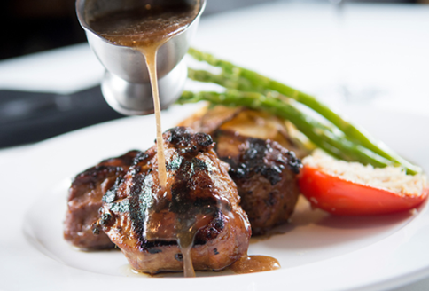 Steak with sauce being poured, asparagus, tomato half, and potatoes on white plate.