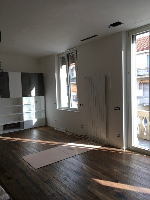 Stanza vuota con pavimento in legno scuro, pareti bianche e luce del sole che filtra dalle finestre e dalla porta-finestra del balcone.