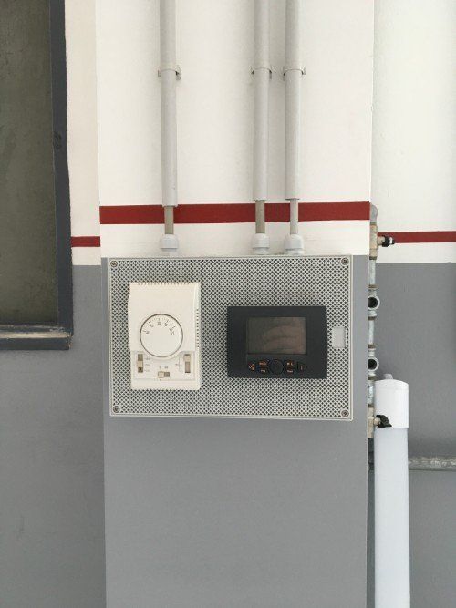 Quadro di controllo a parete con quadranti e display su una parete industriale grigia, con striscia rossa e tubi sovrastanti.