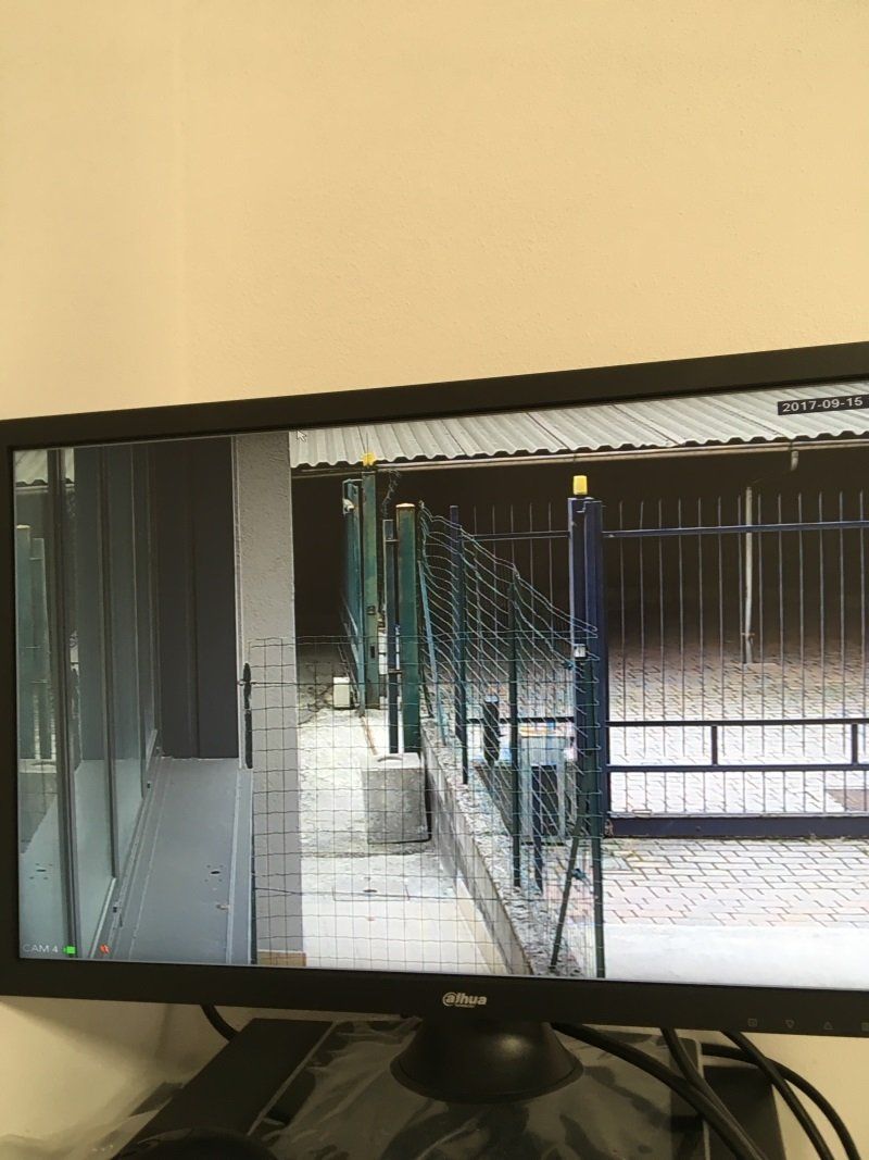 Passaggio pedonale residenziale con recinzione a rete metallica e cancello accanto a un edificio, visto su uno schermo televisivo.