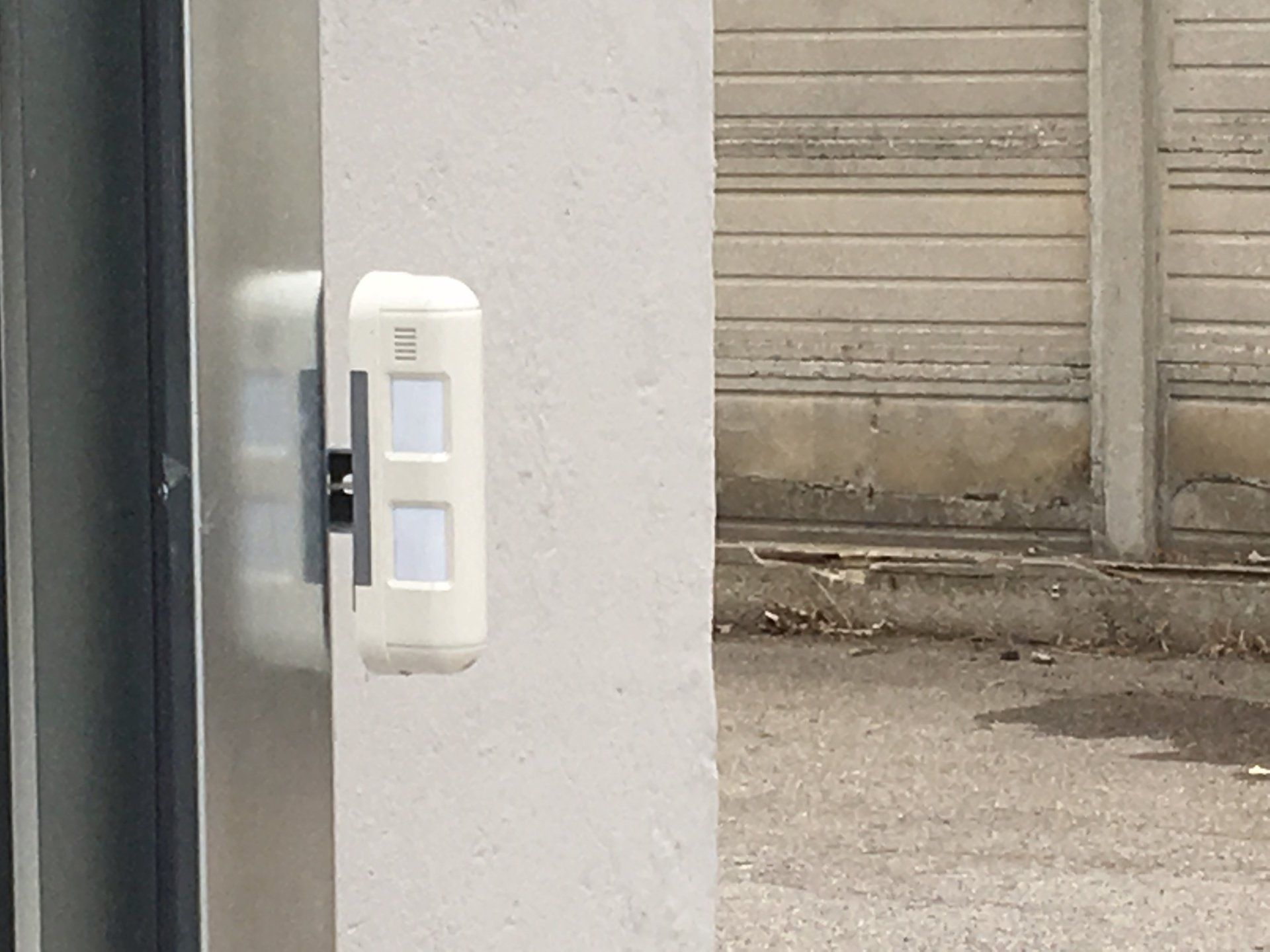 Campanello o pannello citofonico bianco montato a parete accanto a una porta metallica in un vicolo.