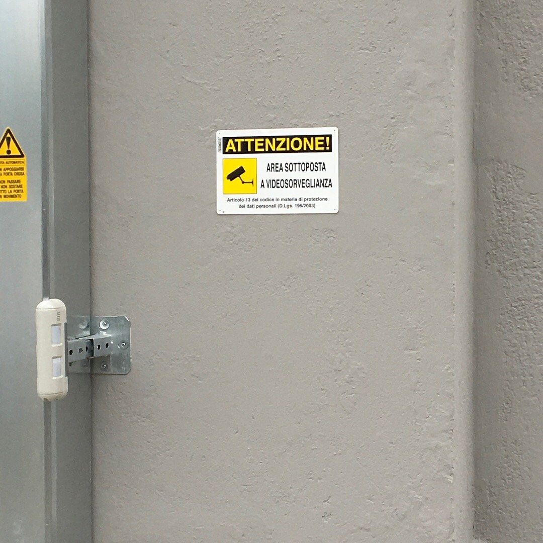 Porta industriale grigia con cartello di avvertimento e maniglia a muro sulla sinistra.