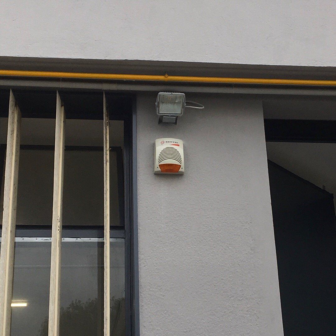 Parete esterna con telecamera di sicurezza e centralina d'allarme montata accanto a una finestra con sbarre.