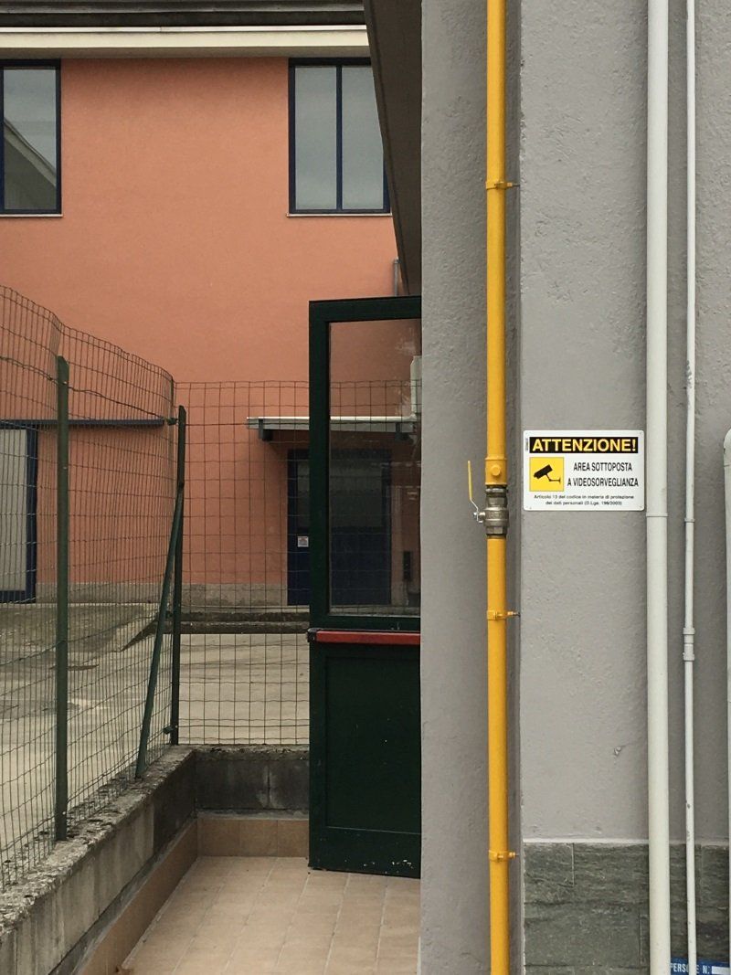 Stretto vicolo accanto a un edificio arancione, con un cancello verde, una recinzione metallica e un tubo giallo su un muro grigio.
