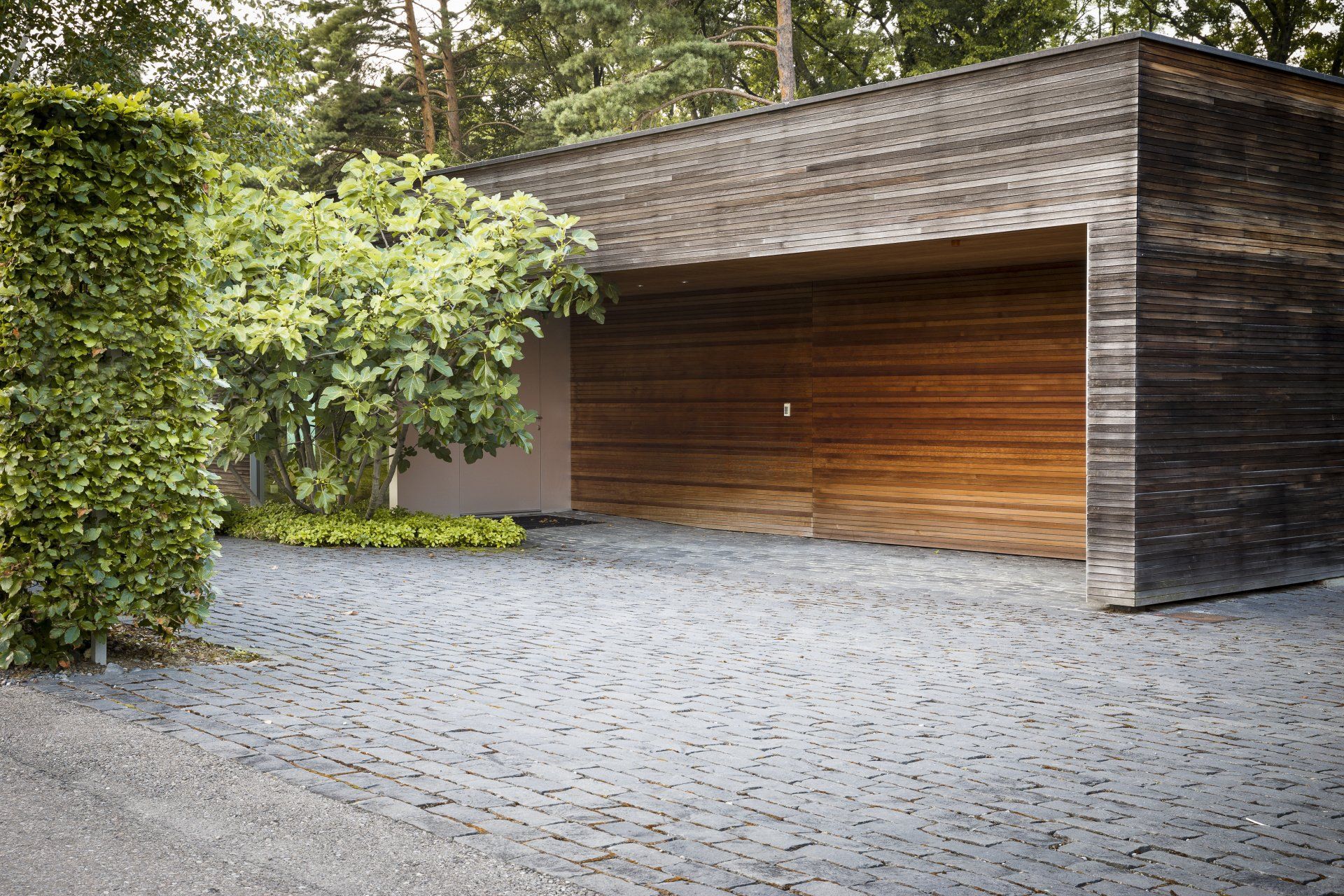 A garage with a wooden door is surrounded by trees and gravel.