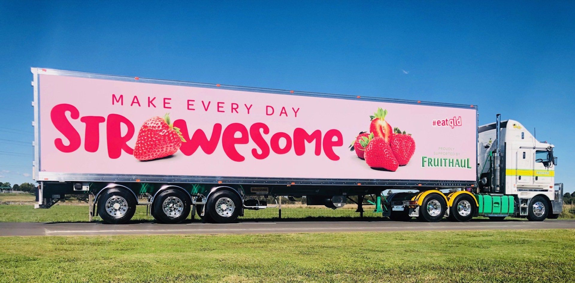A semi-truck trailer with a pink strawberry advertisement on a highway on a sunny day.