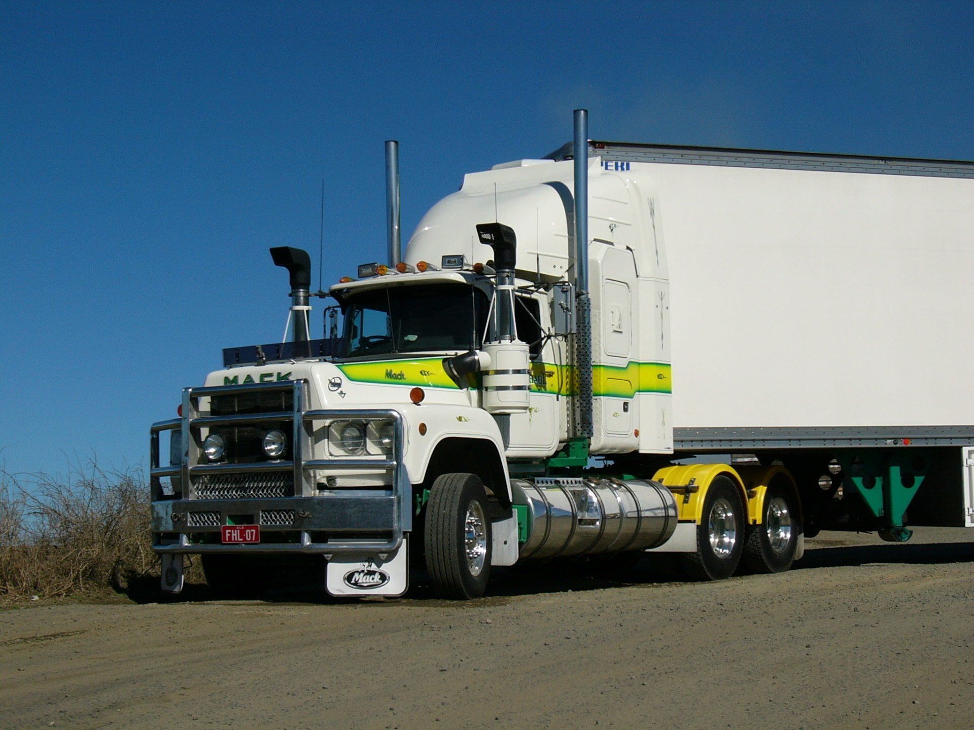 White Mack truck with trailer, on a dirt road, against a blue sky.