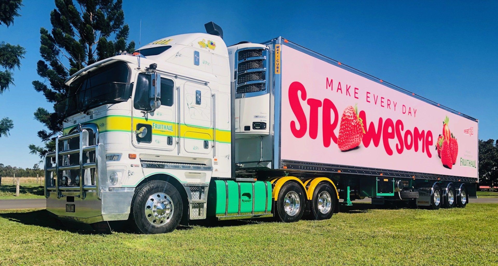 White semi-truck with a trailer advertising strawberries, parked on grass. Trailer says