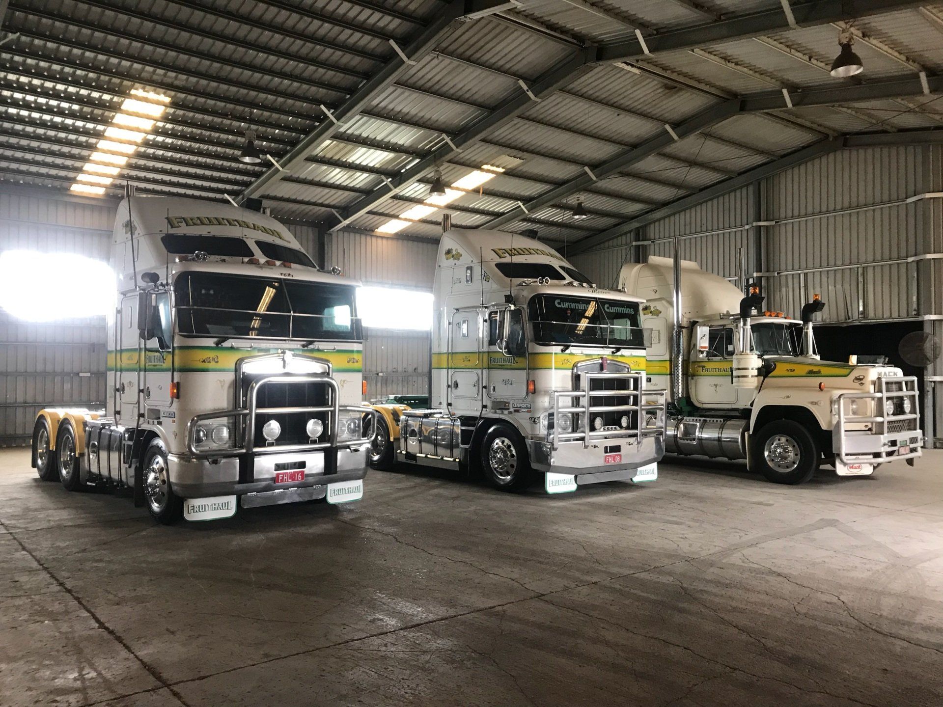 Three semi-trucks parked inside a large garage. The trucks are silver, white, and yellow with chrome details.