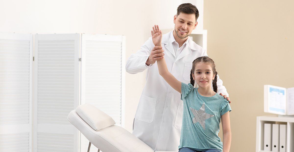 Doctor examining a girl's arm, indoors. The girl is smiling and raising her arm. The doctor wears a white coat.