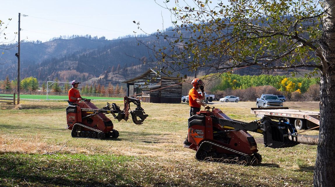 Two men are riding tractors in a field with mountains in the background.