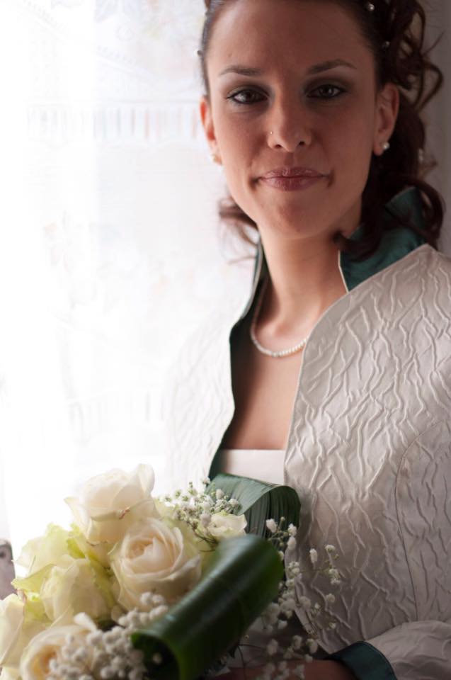 Sposa in abito bianco, con in mano delle rose, che guarda verso la telecamera. Indossa una giacca con finiture verdi vicino a una finestra.