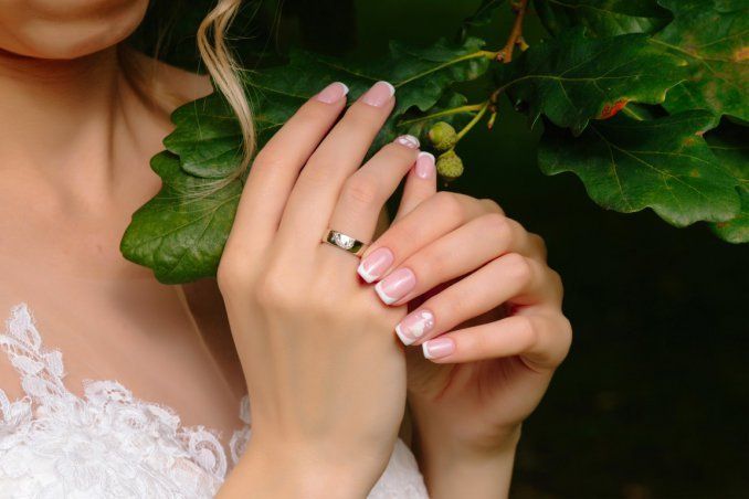 Mani di donna con french manicure e fede nuziale, che tengono in mano foglie verdi, e indossa un abito di pizzo bianco.