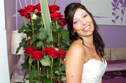 Una donna con un abito bianco senza spalline sorride, accanto a un vaso di rose rosse, in una stanza viola.
