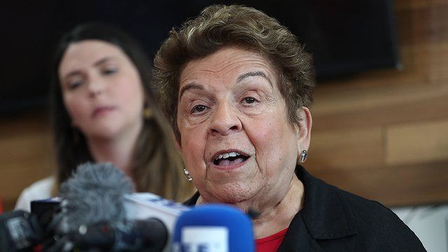 An elderly woman is talking into a microphone with a woman in the background.