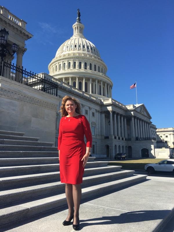 Diana Parr StMarie, Founder of Cyber Instincts™ wearing a red dress stands on the steps of the United States Capitol building under a clear blue sky.
