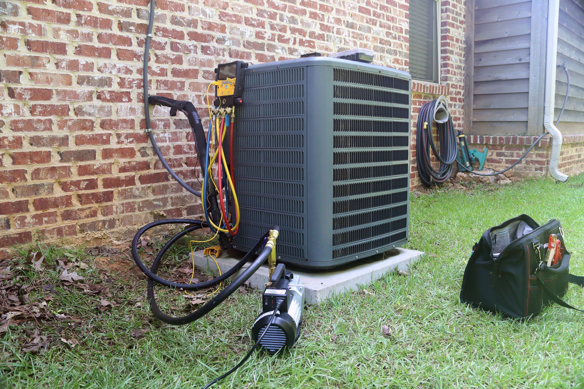HVAC unit being serviced outdoors, brick wall background, hoses and tools present.