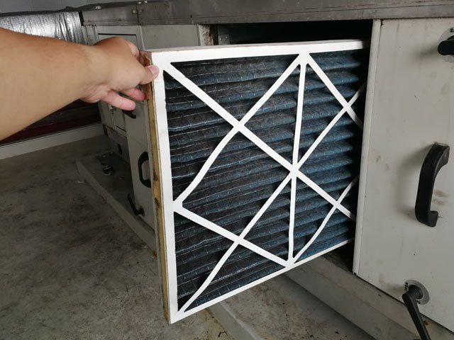 Hand holding a dirty air filter from an HVAC unit. Filter is black with a white frame and supporting lines.