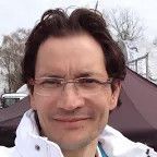 Een man met een bril en een wit jasje lacht naar de camera.