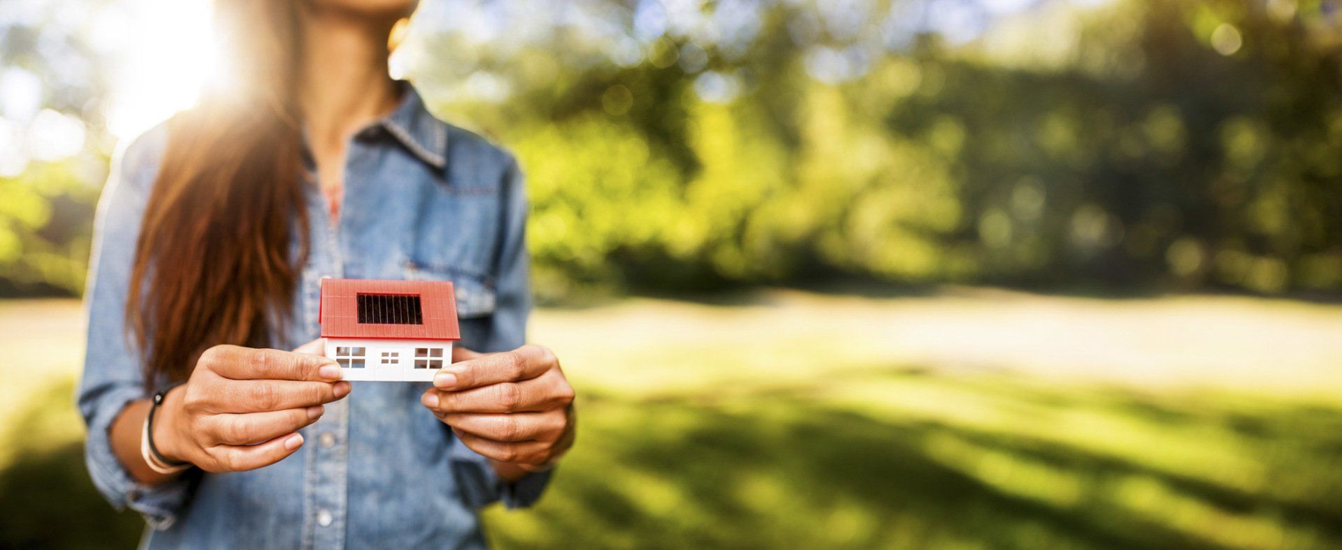 Woman presenting a model of a toy house with solar cells and a sunlit lawn behind her