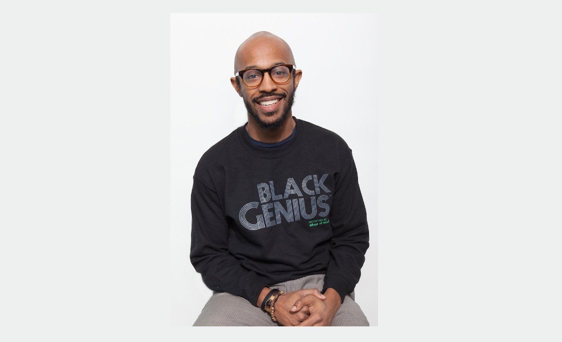 William Jackson, Founder of Village of Wisdom, a Black youth organization empowering their Black genius Will Jackson sits on a stool posing for a photo shoot with white background and black tshirt with slogan Black Genius protected by Village of Wisdom