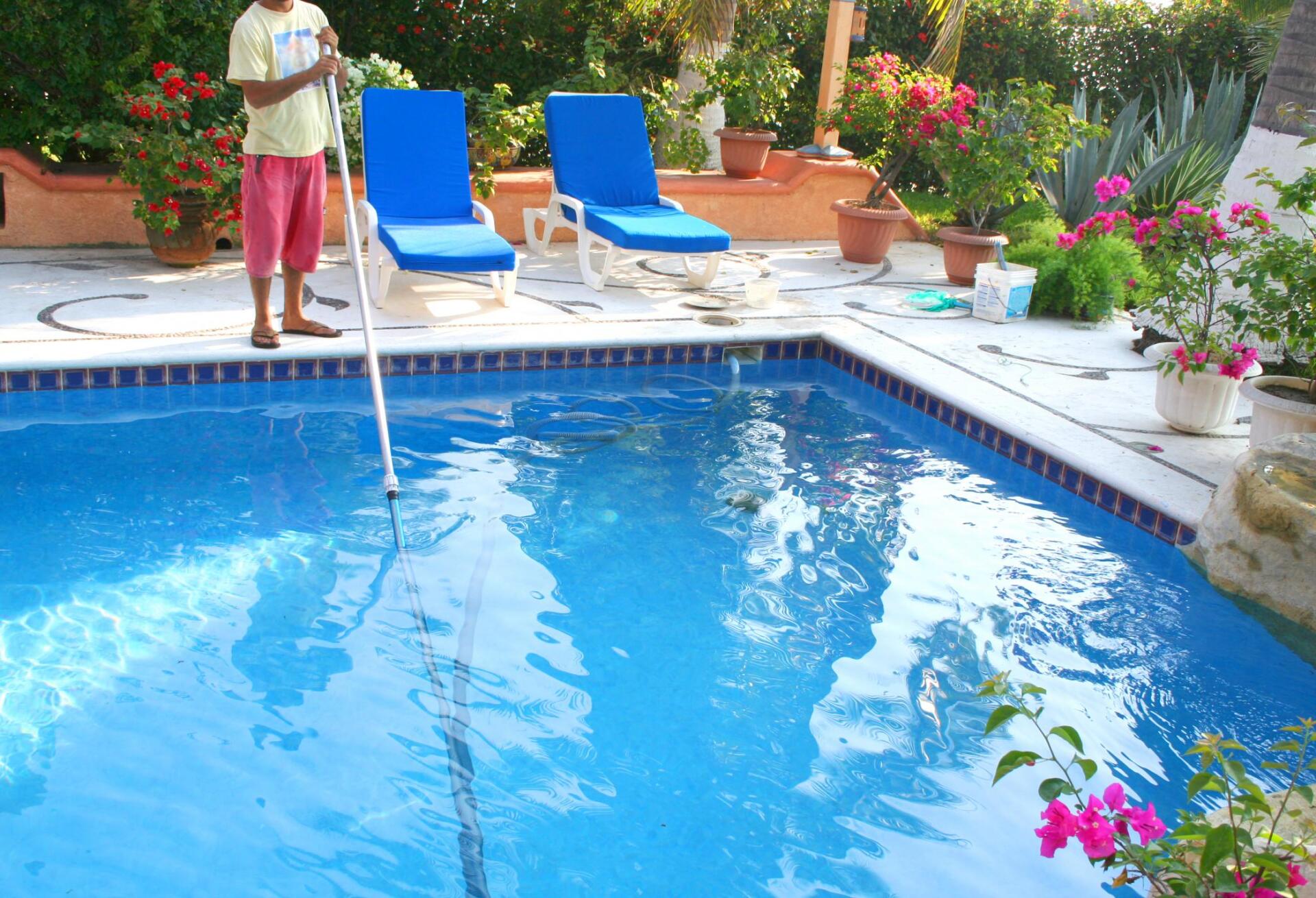 Person cleaning a blue swimming pool with a long-handled tool; two lounge chairs are nearby.