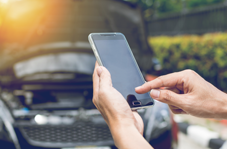 Hands using a smartphone to inspect a car with its hood open outdoors