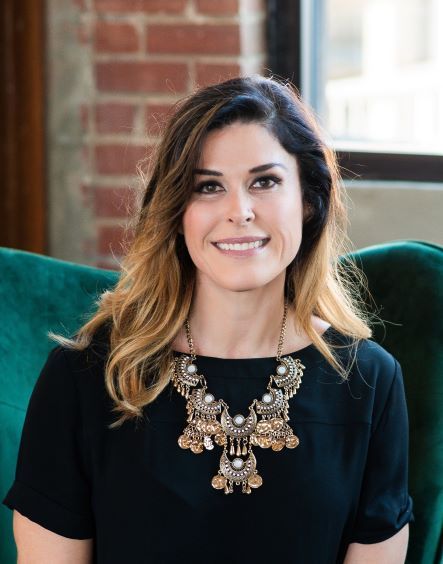 A smiling person with long brown hair, wearing a black top and a large, ornate metal necklace, sitting in a green chair.