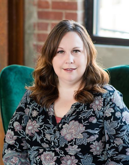 A person with shoulder-length wavy brown hair wears a floral print top while seated in a green chair against a brick wall.