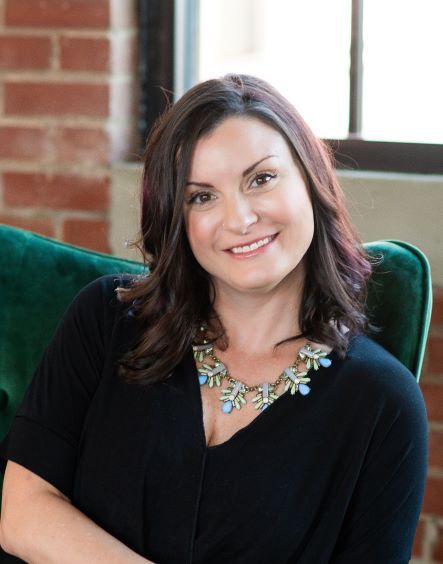A person with dark hair wears a black top and a statement necklace, sitting in a green chair against a brick wall.