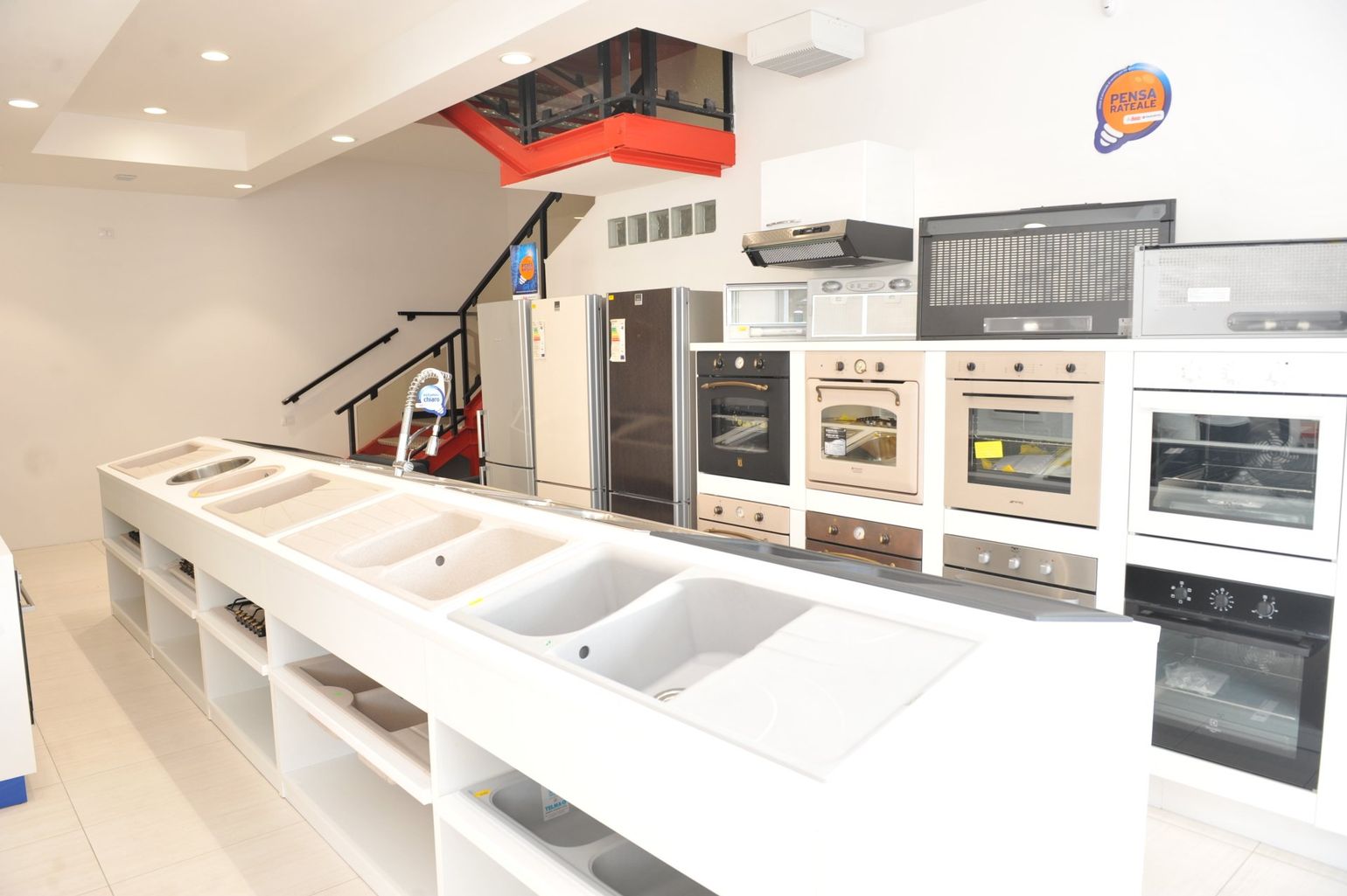 Uno showroom che espone elettrodomestici da cucina, tra cui lavelli, forni e frigoriferi, disposti su scaffali e banconi bianchi.