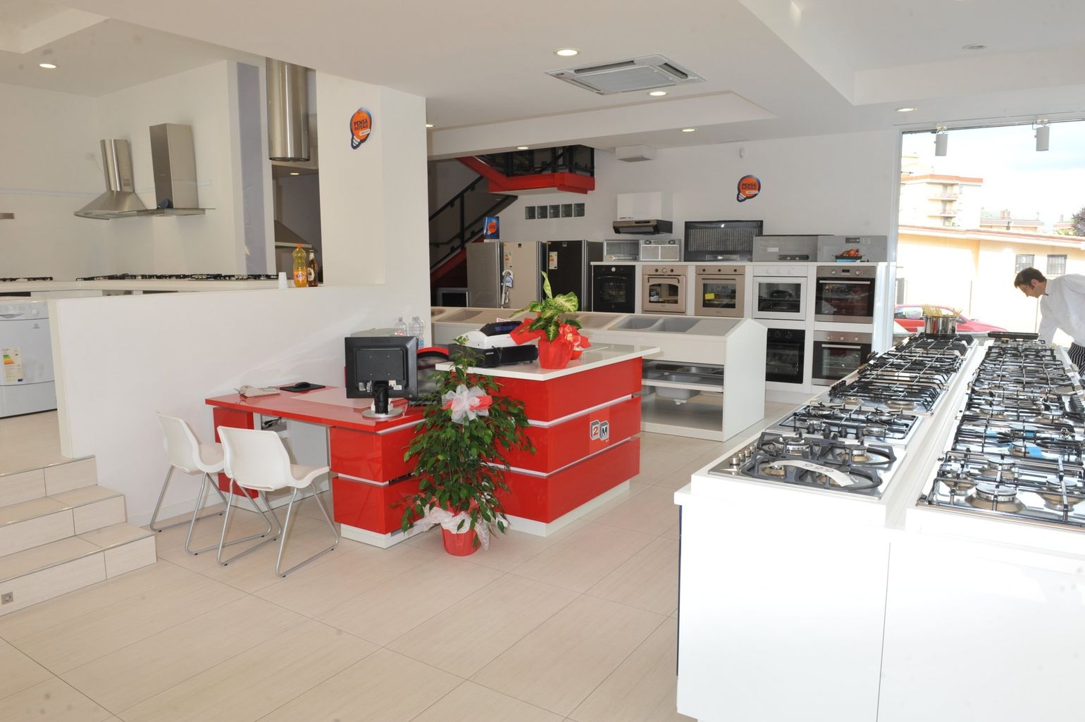 Uno showroom di elettrodomestici con un bancone di vendita rosso, scaffali espositivi con forni a incasso e file di piani cottura in vendita.