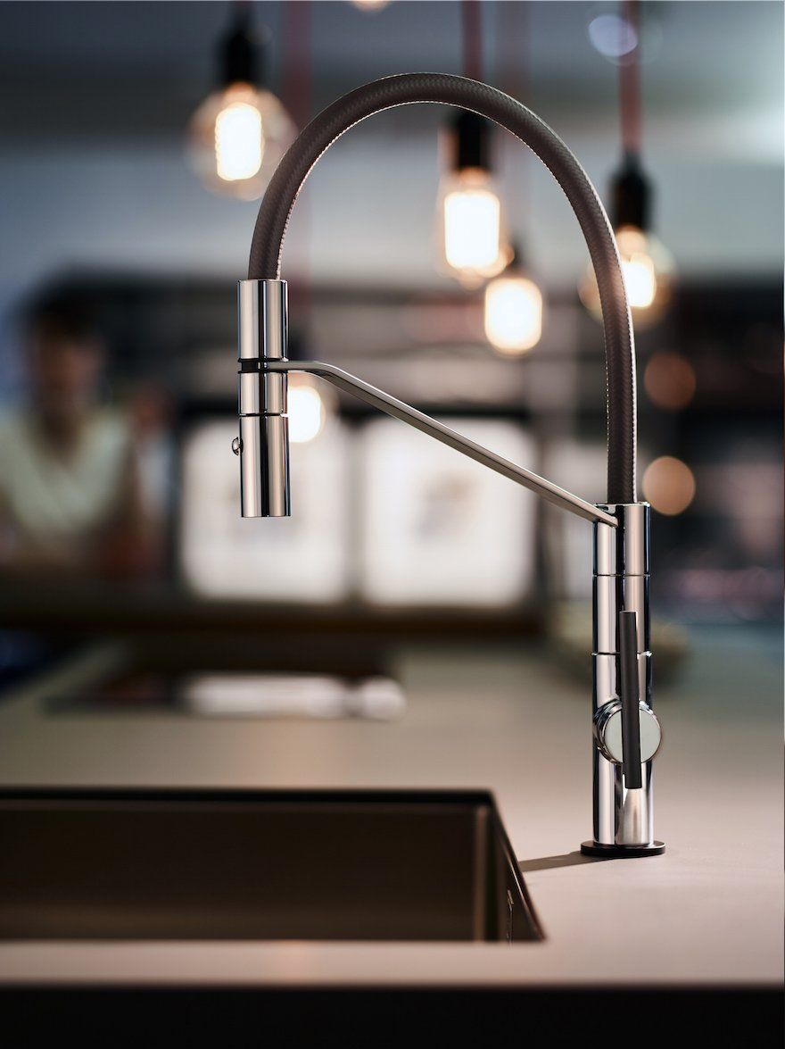 Un moderno rubinetto da cucina estraibile in metallo con un tubo flessibile scuro, sullo sfondo di una cucina sfocata e illuminata da una luce soffusa.