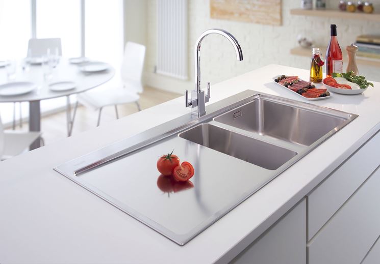 Un moderno lavello da cucina in acciaio inox con zona di taglio e rubinetto, incastonato in un piano di lavoro bianco con alimenti nelle vicinanze.