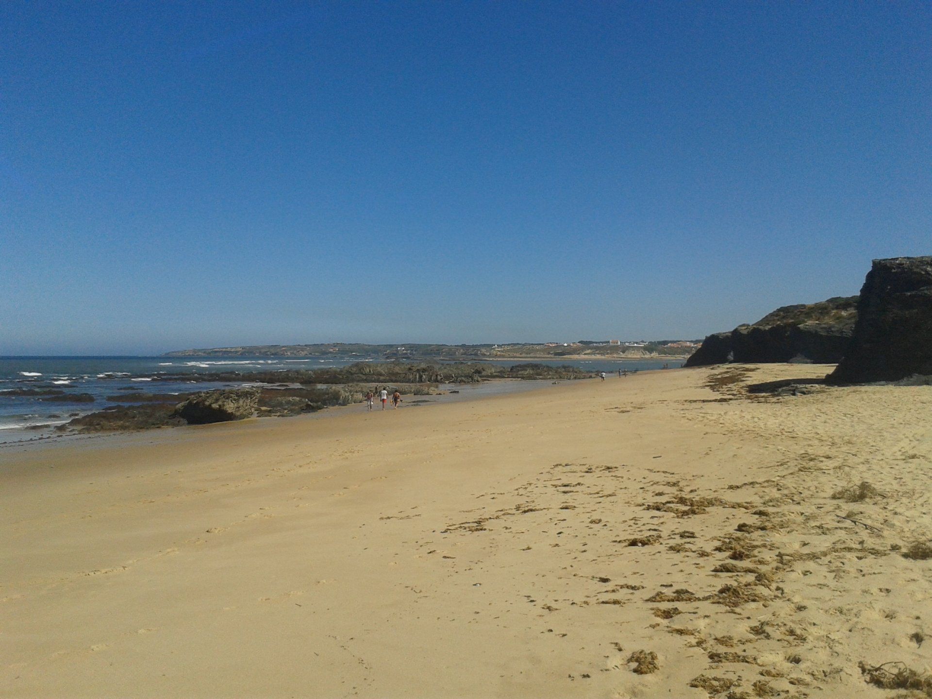Zonnig zandstrand met een kalme oceaan, rotsachtige kustlijn en een heldere blauwe hemel.