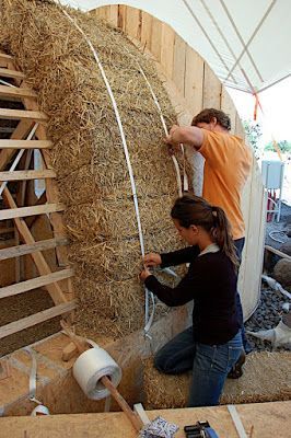 Twee personen bevestigen stro-isolatie met riemen en planken aan een gebogen constructie in een tentvormige werkruimte.